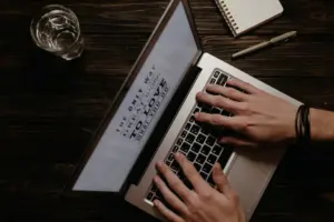A person writing content on a laptop with notes beside it
